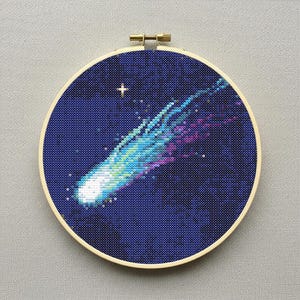 May include: A cross-stitch embroidery of a comet against a dark blue starry background. The comet is depicted with shades of blue, green, and purple, with a bright white head. The embroidery is set in a round wooden hoop.