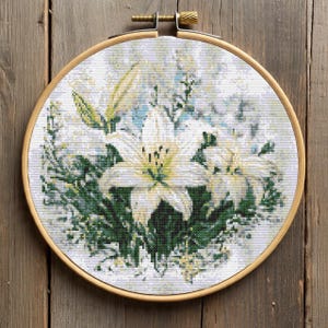 May include: A cross-stitch embroidery of white lilies with green foliage, set in a wooden hoop. The flowers have yellow centres and are surrounded by green leaves and a light blue background. The hoop is made of light-coloured wood.