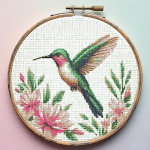 Op de afbeelding: Kruissteekborduurwerk van een kolibrie in vlucht, geplaatst in een houten hoepel. De vogel heeft groene, roze en bruine tinten, met uitgespreide vleugels. Roze en groene bloemelementen omringen de kolibrie en creëren een kunstwerk met een natuurthema.