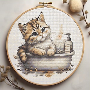 Puede incluir: Un patrón de punto de cruz de un lindo gatito sentado en una bañera con una botella de jabón. El gatito es marrón y blanco con grandes ojos azules. La bañera es blanca con un borde azul. La botella de jabón es blanca con una etiqueta azul.