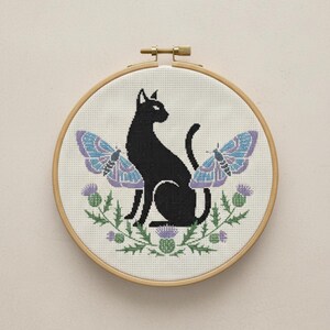 May include: Cross-stitch embroidery in a wooden hoop. The design features a black cat, two blue and purple butterflies, and green thistles. The embroidery is on a cream-colored fabric.