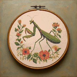 Puede incluir: Bordado de punto de cruz de una mantis religiosa verde rodeada de flores naranjas y hojas verdes. El bordado está colocado en un aro de madera, sobre un fondo claro. El aro está montado en un marco de madera de color verde azulado.