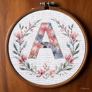May include: Cross-stitch embroidery in a wooden hoop. The design features the letter "A" in a floral pattern of pink and blue hues, surrounded by a wreath of pink flowers and green leaves. The background is white.