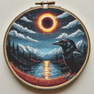 May include: Cross-stitch art in a wooden hoop depicts a dark raven perched before a landscape. The scene includes a river, mountains, and a celestial event with a bright orange and red sun. The sky is filled with clouds.