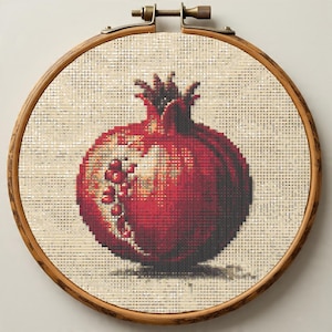 May include: A cross-stitch embroidery of a pomegranate fruit, displayed in a wooden hoop. The fruit is rendered in shades of red, with visible seeds and a textured appearance. The background is a neutral beige, and the hoop has a brass adjustment mechanism.