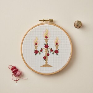 Könnte beinhalten: Kreuzstich-Stickerei eines Kerzenständers mit drei Kerzen und roten Rosen, in einem Holzreifen. Die Kerzen haben gelbe und rosa Flammen. Ein goldener Fingerhut und rotes und rosa Garn sind ebenfalls zu sehen.