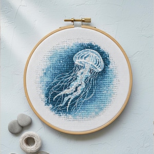 May include: A cross-stitch embroidery of a jellyfish in a wooden hoop. The jellyfish is white and light blue against a dark blue background. The hoop is made of light-colored wood. The image includes a spool of thread and two small gray stones.