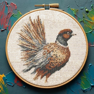 May include: A cross-stitch embroidery of a pheasant in a wooden hoop. The bird features brown, gray, and white details, with a detailed wing spread. The background is a neutral color, and the hoop is set against a dark teal surface with colorful paint strokes.