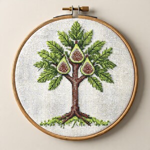 Può includere: Ricamo a punto croce di un albero di fico, con un tronco marrone, foglie verdi e tre fichi. Il ricamo è posto in un telaio di legno, mostrando un artigianato dettagliato e fatto a mano. Il disegno è su un tessuto bianco.
