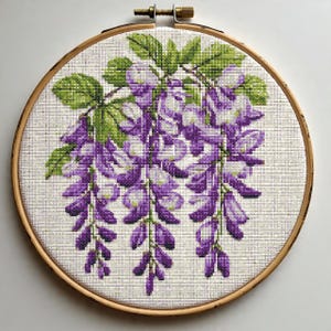 May include: A cross-stitch embroidery of wisteria flowers in a wooden hoop. The design features cascading purple and white blossoms with green leaves. The embroidery is set against a neutral fabric background, creating a handcrafted floral artwork.