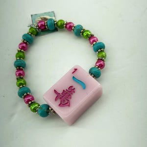 Könnte beinhalten: Ein farbenfrohes Perlenarmband mit einer rosa Mahjong-Kachel mit chinesischen Schriftzeichen und einem blauen Akzent. Das Armband ist mit türkisfarbenen, grünen und rosa Perlen aufgereiht, mit silberfarbenen Abstandshaltern. Ein einzigartiges Accessoire.