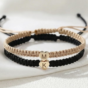 Könnte beinhalten: Zwei passende Freundschaftsarmbänder. Eines ist schwarz, das andere ist beige. Jedes Armband hat einen goldenen Herzanhänger mit den Initialen "M" und "K". Die Armbänder sind aus geflochtenem Kordel gefertigt und werden auf einer weißen Oberfläche präsentiert.
