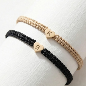 Op de afbeelding: Twee macramé armbanden, één zwart en één beige, elk met een gouden hartbedel. De zwarte armband heeft een gouden hart met de letter "B", en de beige armband heeft een gouden hart met de letter "K".