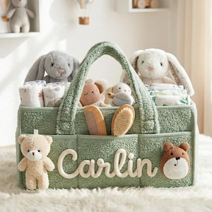 May include: A sage green baby caddy with the name "Carlin" embroidered on the front. The caddy is filled with baby essentials, including plush bunnies, diapers, and grooming tools. A teddy bear and a bear face are attached to the caddy.