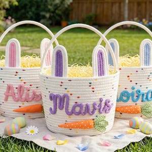 以下が含まれることがあります： ウサギの耳、パーソナライズされた名前、ニンジンのアクセントが付いた、白いロープのイースターバスケットが3つ。各バスケットにはハンドルがあり、黄色のフィラーが詰められています。カラフルな卵と花が白い布の上にバスケットを囲んでいます。