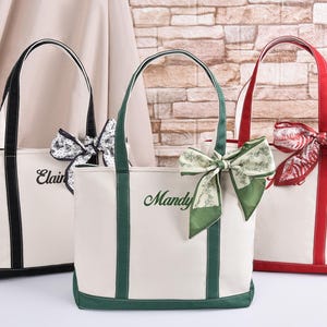 May include: Three canvas tote bags with contrasting trim and handles. One bag has black trim, another green, and the third red. Each bag has a decorative bow and a name embroidered on the front.