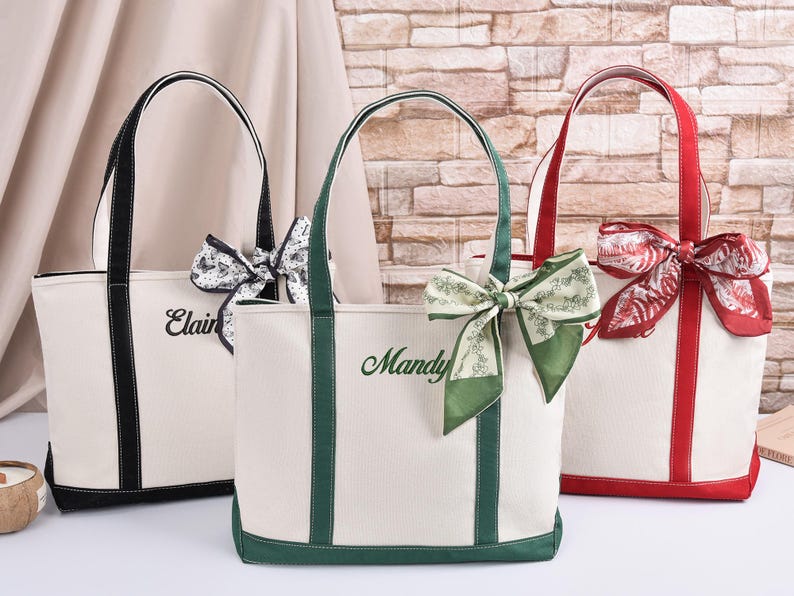 May include: Three canvas tote bags with contrasting trim and handles. One bag has black trim, another green, and the third red. Each bag has a decorative bow and a name embroidered on the front.