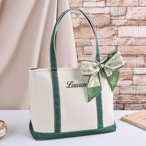 May include: A cream canvas tote bag with green trim and handles. The word "Lauren" is embroidered in green script. A green and white patterned bow is attached to the handle. A coconut shell candle and a book are also visible.