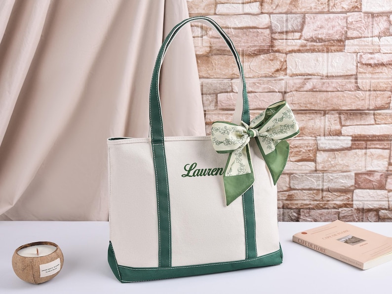 May include: A cream canvas tote bag with green trim and handles. The word "Lauren" is embroidered in green script. A green and white patterned bow is attached to the handle. A coconut shell candle and a book are also visible.