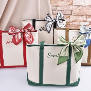 May include: Five personalised canvas tote bags with colourful accents and decorative bows. Each bag features a different colour trim and bow, with names embroidered on the front. The bags are cream-coloured with red, green, black, and brown accents.