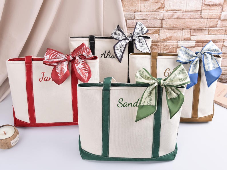 May include: Five personalised canvas tote bags with colourful accents and decorative bows. Each bag features a different colour trim and bow, with names embroidered on the front. The bags are cream-coloured with red, green, black, and brown accents.