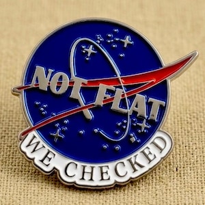 May include: A round, blue and silver pin with the text "NOT FLAT" and "WE CHECKED". The pin features a red swoosh and white stars, resembling a NASA-inspired design. The pin is on a tan, textured surface.