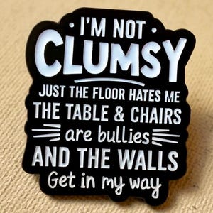 May include: A black and white enamel pin with the text "I'M NOT CLUMSY" and other humorous phrases about clumsiness. The pin is rectangular with rounded edges and is set against a tan background.