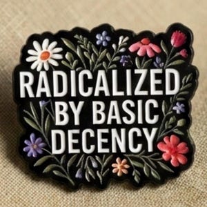 May include: Black pin with the text "RADICALIZED BY BASIC DECENCY" in white. The pin is decorated with colorful flowers and leaves. The pin is on a tan fabric surface.