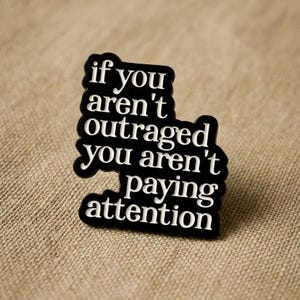 May include: A black and white pin with the quote "if you aren't outraged you aren't paying attention." The pin has a black background with white text and a white border. The pin is on a tan, textured surface.