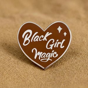 May include: A heart-shaped enamel pin with a brown background and a silver border. The pin features the words "Black Girl Magic" in white cursive lettering and two small white starbursts.