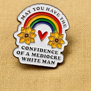 Op de afbeelding: Witte emaille pin met een regenboog, twee gele zonnebloemen en een rood hart. De pin heeft de tekst "MAY YOU HAVE THE CONFIDENCE OF A MEDIOCRE WHITE MAN" in zwarte letters. De pin ligt op een bruin textiel oppervlak.