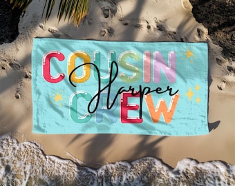 May include: A light blue beach towel with the words "COUSIN CREW Harper" in colorful bubble letters. The towel is on a sandy beach with ocean waves. The text "FREE STORAGE BAG" is at the top.