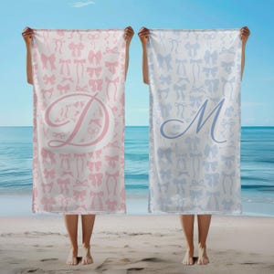 Könnte beinhalten: Zwei Strandtücher, eines rosa und eines hellblau, werden vor einem Strand und Ozean gehalten. Das rosafarbene Handtuch hat einen großen Buchstaben "D" und ein Muster aus rosa Schleifen. Das hellblaue Handtuch hat einen großen Buchstaben "M" und ein Muster aus hellblauen Schleifen.