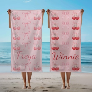 May include: Two pink beach towels with a repeating pattern of cherries and bows. One towel has the name "Freya" in a script font, and the other has "Winnie". The towels are held up on a beach.