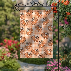 May include: A garden flag with a repeating pattern of orange pumpkins with white detailing on an orange and white plaid background. The flag is suspended from a black metal garden stake, ideal for autumn decoration.