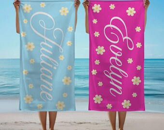 May include: Two beach towels are held up, one light blue and one magenta. Both towels have a floral pattern with white daisies and the names "Juliane" and "Evelyn" in cursive. The text "FREE STORAGE BAG" is at the top.