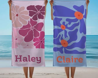 May include: Two beach towels with floral designs are held up on a beach. One towel is light pink with pink flowers and the name "Haley". The other is blue with purple flowers and the name "Claire".