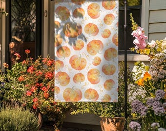 May include: A white garden flag with a repeating pattern of watercolour pumpkins in orange and yellow hues. The pumpkins vary in size and are decorated with small, curly lines. The flag is displayed outdoors, surrounded by plants and flowers.