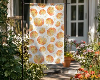 May include: A garden flag with a repeating pattern of orange and yellow watercolor pumpkins with green stems and curly vines on a white background. The flag is displayed outdoors.