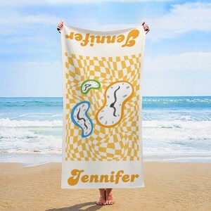 Könnte beinhalten: Ein weißes Strandtuch mit einem gelb-weiß karierten Muster und Designs mit schmelzenden Uhren. Das Wort "Jennifer" ist oben und unten auf dem Handtuch in Orange aufgedruckt. Das Handtuch wird an einem Strand mit dem Meer im Hintergrund gehalten.