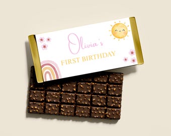 Könnte beinhalten: Ein Schokoriegel mit dekorativer Verpackung. Die Verpackung zeigt den Text "Olivia's FIRST BIRTHDAY" in Rosa und Gold, zusammen mit einer lächelnden Sonne, einem Regenbogen und floralen Akzenten. Der Schokoriegel ist dunkelbraun und hat eine strukturierte Oberfläche.