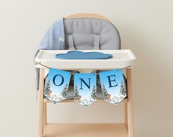 May include: A blue banner with the word "ONE" in navy blue letters is strung across a high chair. The banner has a winter scene with trees and animals. The high chair has a gray seat and a blue cloud-shaped tray.