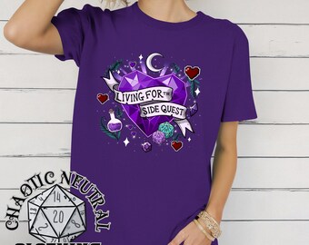 May include: A purple t-shirt featuring a graphic design with a purple heart, dice, and the text "LIVING FOR THE SIDE QUEST." The shirt also has the text "CHAOTIC NEUTRAL CLOTHING" on the bottom left.