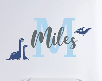 Może przedstawiać: Naklejka ścienna z niebieskim i szarym dinozaurem z imieniem "Miles" w jasnoniebieskiej literze M. Naklejka znajduje się na białej ścianie nad łóżkiem z niebieskimi i szarymi poduszkami.