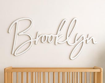 Może przedstawiać: Scena pokoju dziecięcego z drewnianym łóżeczkiem z białym materacem, fotelem bujanym i małym stolikiem. Biały znak z napisem "Brooklyn" jest zamontowany na ścianie nad łóżeczkiem. Pleciony puf i roślina doniczkowa dopełniają pokój.