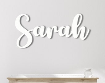May include: White wooden sign with the name "Sarah" written in a cursive font. The sign is mounted on a white wall above a wooden dresser.