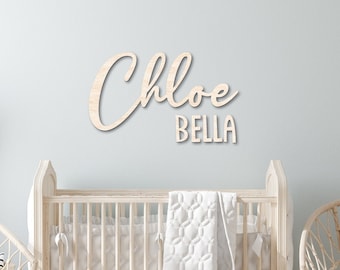 May include: A nursery room with a wooden sign that reads "Chloe Bella" in cursive script. The sign is mounted on a light blue wall. There is a crib with a white blanket and a wicker chair with a white cushion in the room.