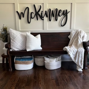 May include: A dark wood bench with a white throw blanket and several white and cream pillows. The bench is decorated with a black wooden sign that reads "McKinney".