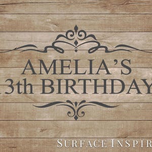 May include: A wooden sign with the text "AMELIA'S 13th BIRTHDAY" in dark gray. The text is centered and framed by decorative flourishes. The words "SURFACE INSPIRED" are at the bottom of the sign.