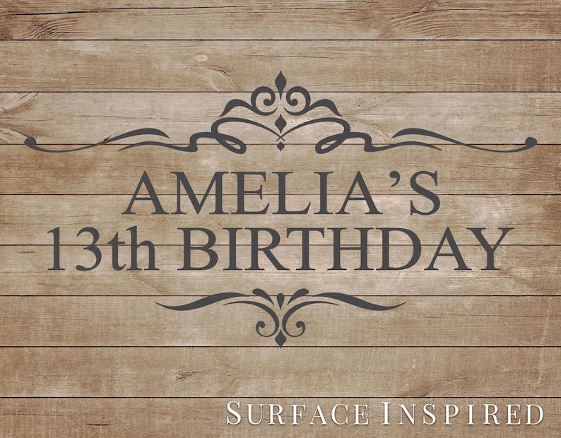 May include: A wooden sign with the text "AMELIA'S 13th BIRTHDAY" in dark gray. The text is centered and framed by decorative flourishes. The words "SURFACE INSPIRED" are at the bottom of the sign.